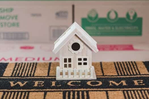 A small decorative wooden house on a welcome mat surrounded by moving boxes indoors.