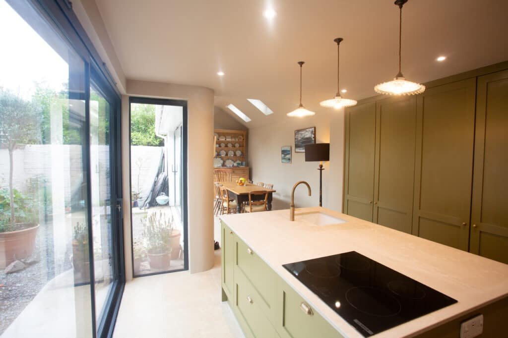 Light-filled kitchen with green island unit, pendant lights, and floor-to-ceiling glazing facing the garden.